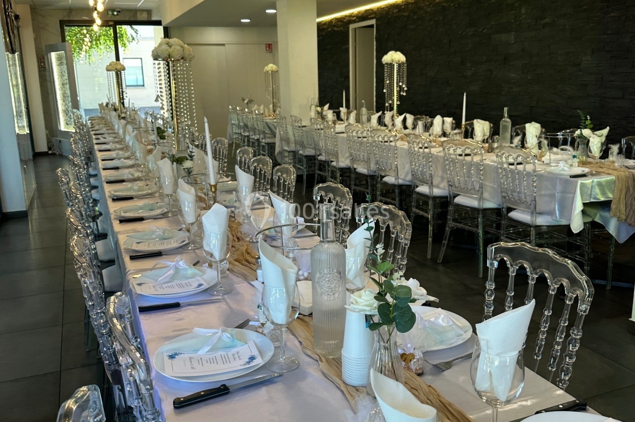 Salle de réception décorée avec de longues tables alignées, nappes blanches, chaises transparentes et centres de table…