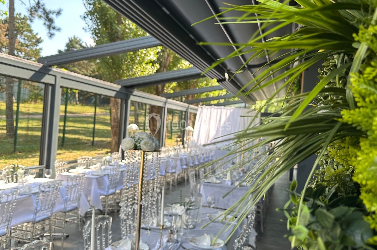 Salle de réception lumineuse avec tables dressées, chaises transparentes et décorations végétales, vue sur un espace…
