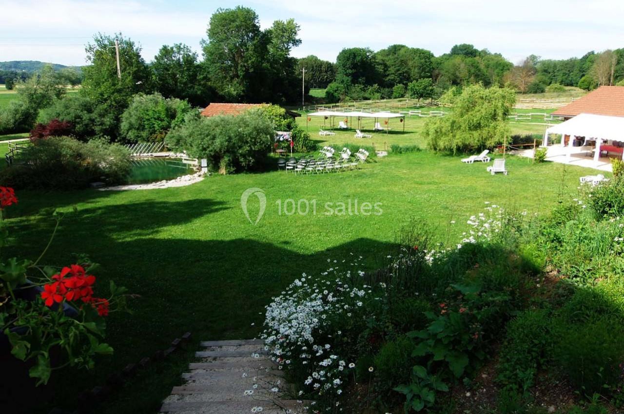 Location salle Ornacieux (Isère) - Domaine du Moulin Piongo #19 Jardin verdoyant avec pelouse, fleurs, chaises disposées en cercle et bâtiments en arrière-plan par temps ensoleillé.