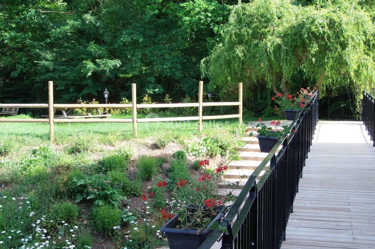 Location salle Ornacieux (Isère) - Domaine du Moulin Piongo #20 Passerelle en bois bordée de jardinières fleuries, menant à un espace vert avec arbres et clôture en bois.