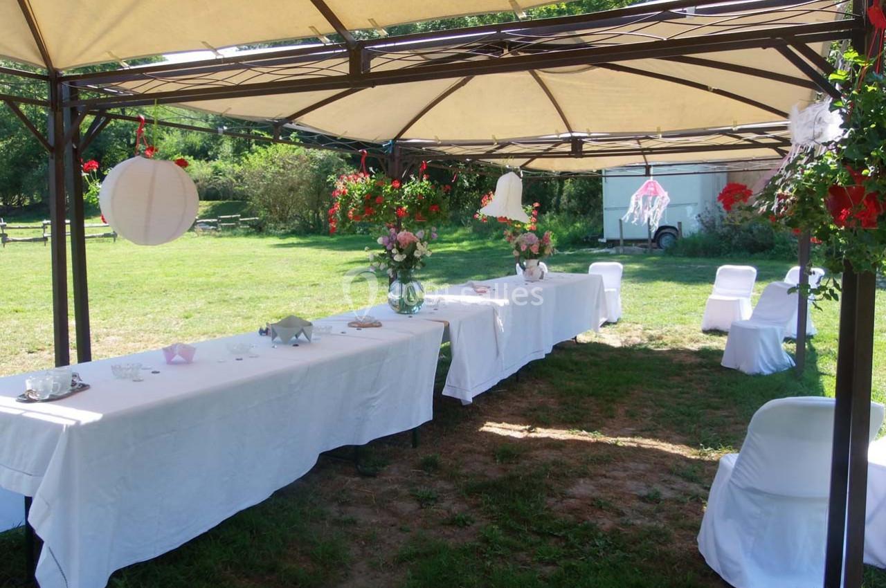Location salle Ornacieux (Isère) - Domaine du Moulin Piongo #17 Tables décorées sous une tonnelle avec nappes blanches, fleurs et lanternes, dans un jardin verdoyant.