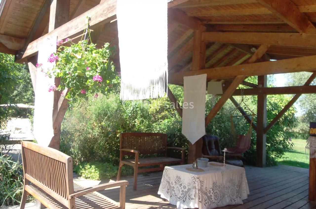 Location salle Ornacieux (Isère) - Domaine du Moulin Piongo #14 Terrasse en bois couverte avec bancs, table décorée d'une nappe blanche et plantes suspendues en fleurs.