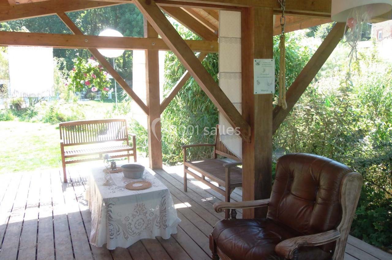 Location salle Ornacieux (Isère) - Domaine du Moulin Piongo #16 Terrasse en bois couverte avec fauteuil, bancs, table décorée et vue sur un jardin verdoyant en arrière-plan.