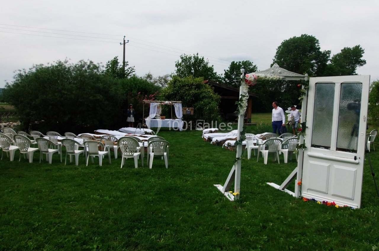 Location salle Ornacieux (Isère) - Domaine du Moulin Piongo #24 Chaises blanches disposées en extérieur sur une pelouse, face à une arche décorée pour une cérémonie.