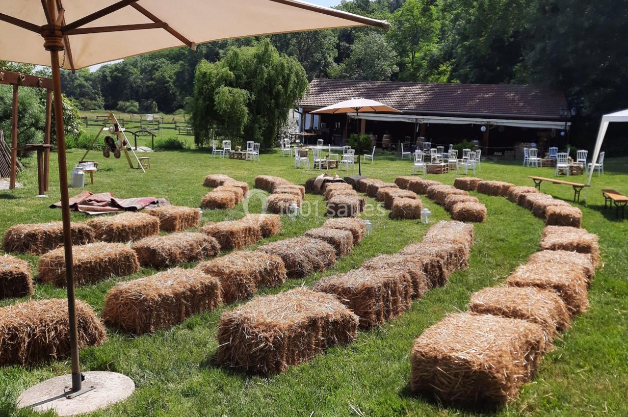 Location salle Ornacieux (Isère) - Domaine du Moulin Piongo #15 Bottes de paille disposées en rangées sur une pelouse, formant un espace assis en plein air près d'un bâtiment et d'arbres.