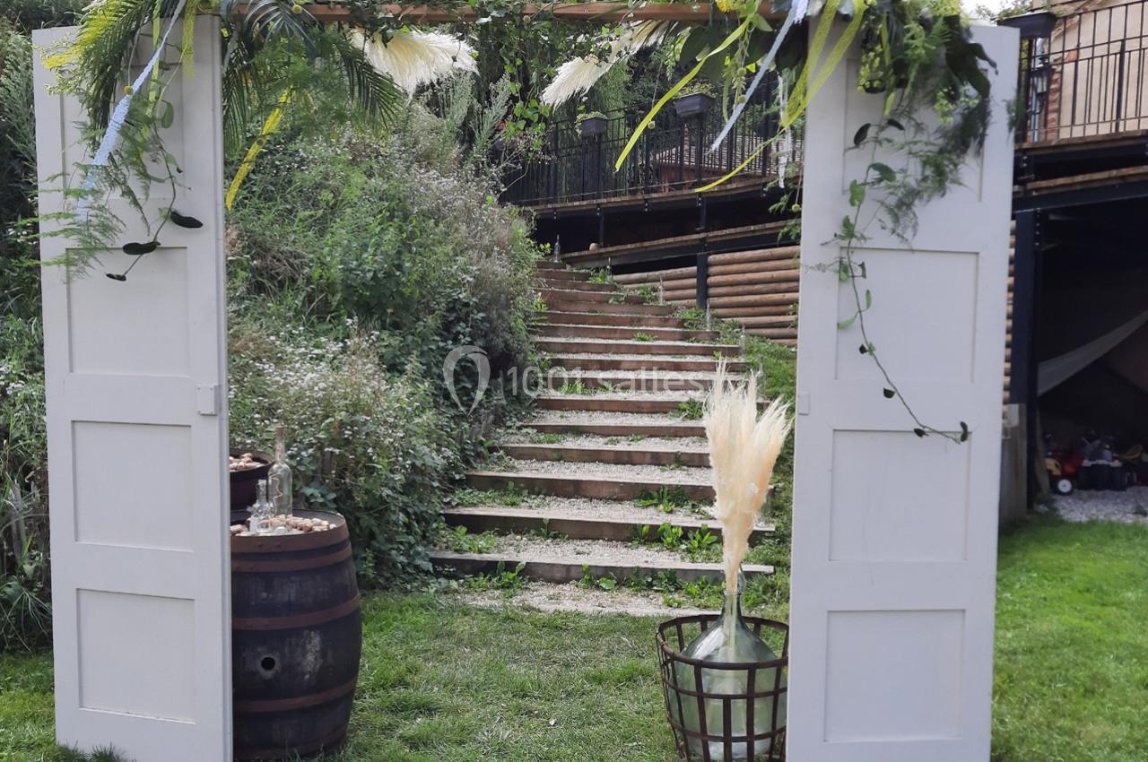 Location salle Ornacieux (Isère) - Domaine du Moulin Piongo #25 Portique décoratif en bois blanc avec feuillages et fleurs, placé devant un escalier extérieur en pierre.