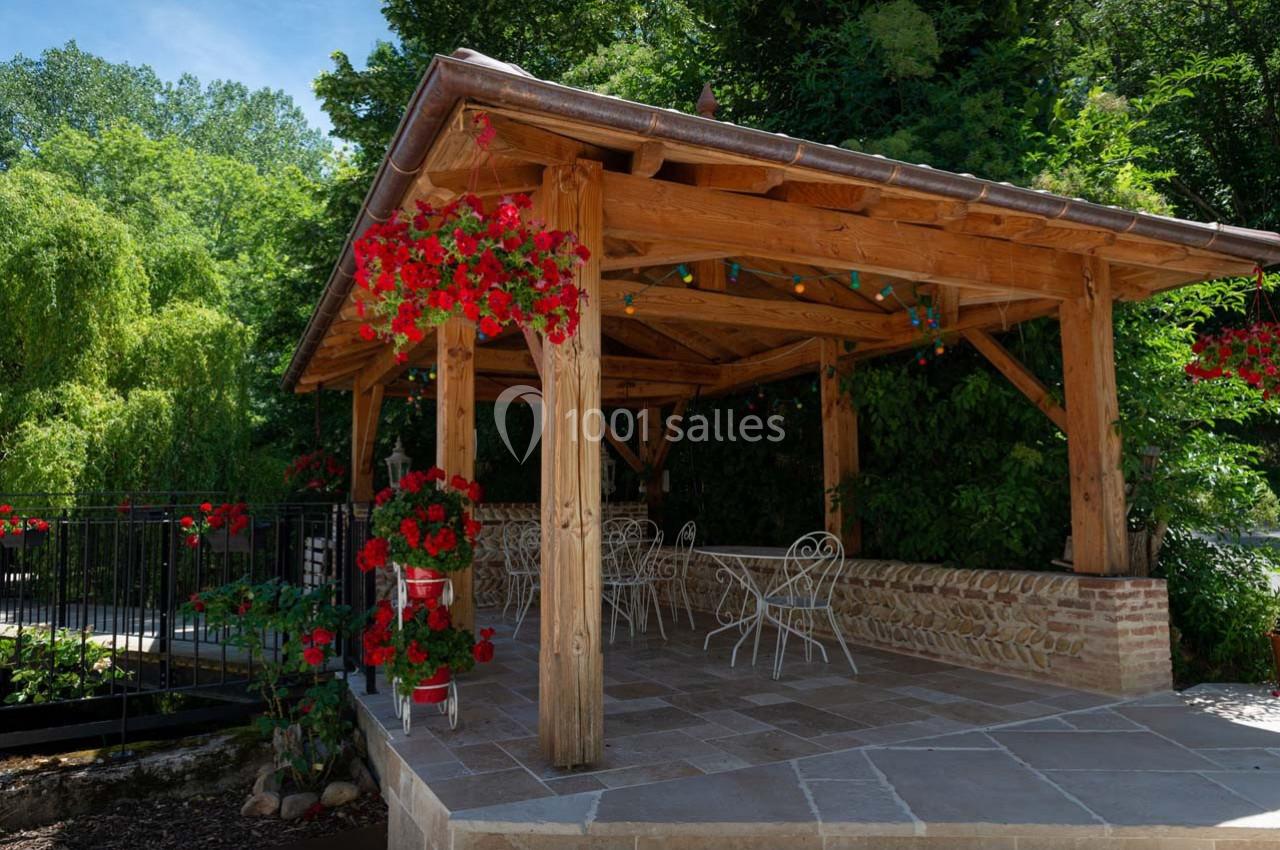 Location salle Ornacieux (Isère) - Domaine du Moulin Piongo #9 Pergola en bois avec des tables et chaises blanches, entourée de fleurs rouges et de verdure.