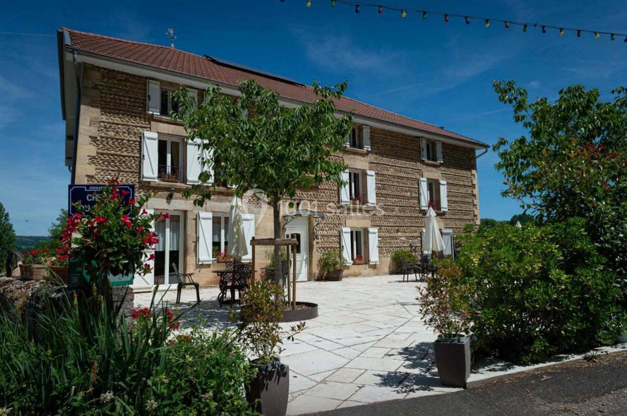 Location salle Ornacieux (Isère) - Domaine du Moulin Piongo #2 Façade d'une maison en pierre avec terrasse fleurie, arbres et ciel bleu en arrière-plan.