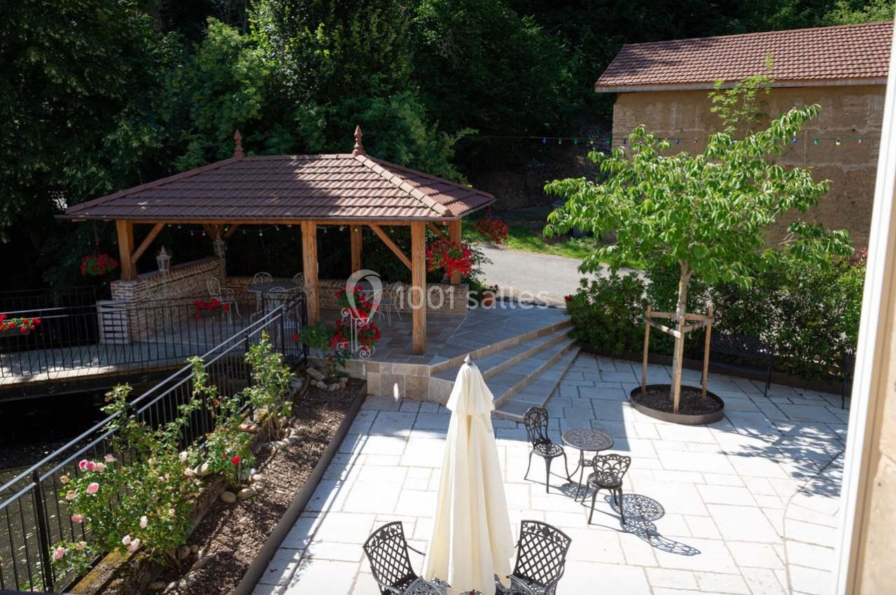 Location salle Ornacieux (Isère) - Domaine du Moulin Piongo #8 Terrasse pavée avec mobilier de jardin, pergola en bois, parterre fleuri et arbres, entourée de verdure.