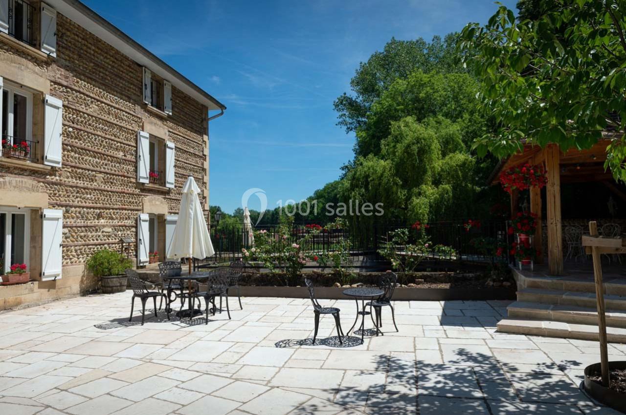 Location salle Ornacieux (Isère) - Domaine du Moulin Piongo #7 Terrasse pavée avec tables et chaises en fer forgé, entourée de verdure et d'un bâtiment en pierre avec volets blancs.