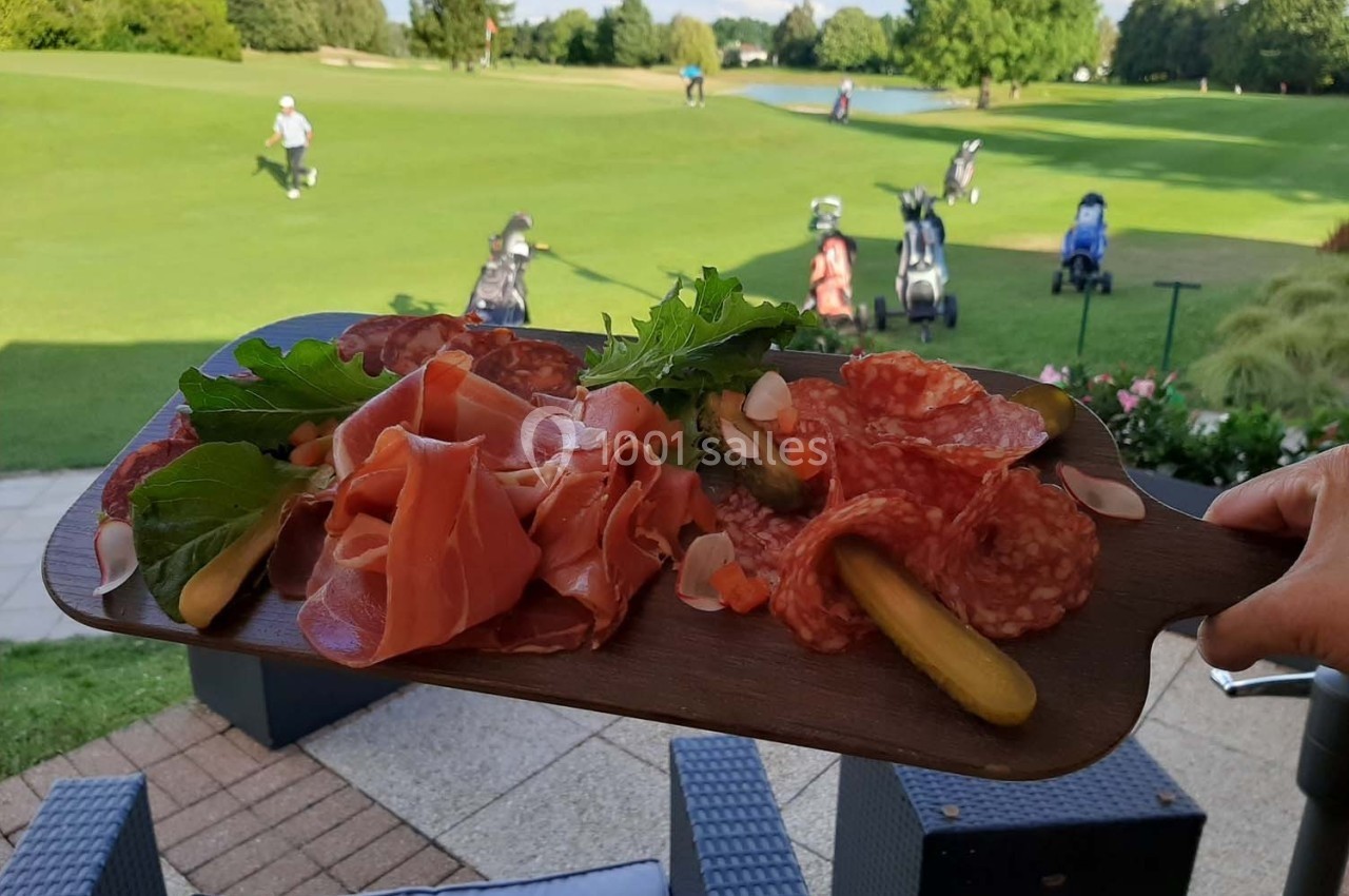 Plateau de charcuterie avec cornichons et salade, tenu en extérieur près d'un terrain de golf.