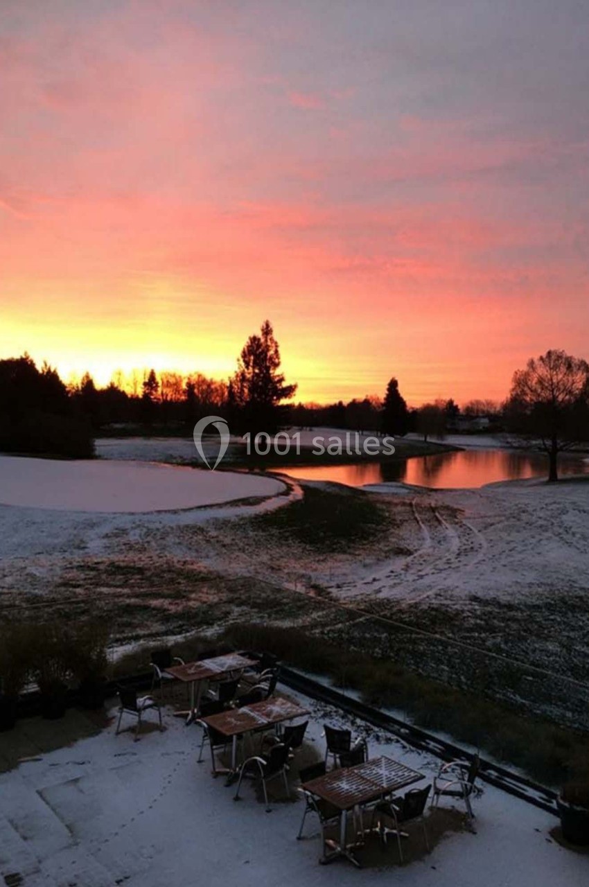 Paysage hivernal avec un terrain enneigé, des tables extérieures et un lac sous un ciel de coucher de soleil coloré.