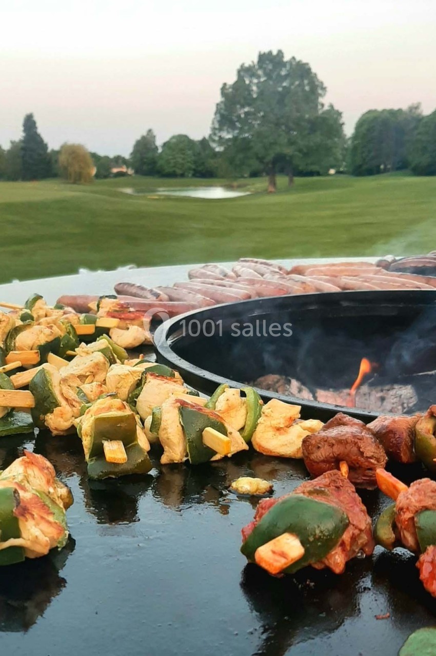 Brochettes de légumes et de viande grillant sur une plancha ronde avec un paysage verdoyant en arrière-plan.