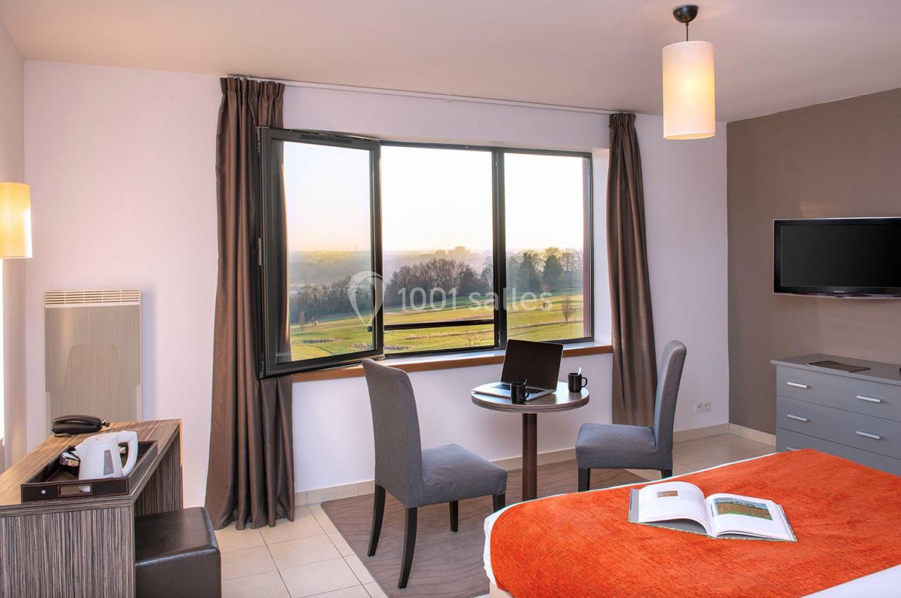 Chambre lumineuse avec lit, table ronde, deux chaises, fenêtre donnant sur un paysage, et télévision murale.