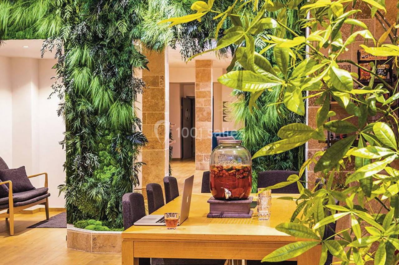 Espace intérieur lumineux avec table en bois, plantes vertes abondantes et fauteuils en arrière-plan.