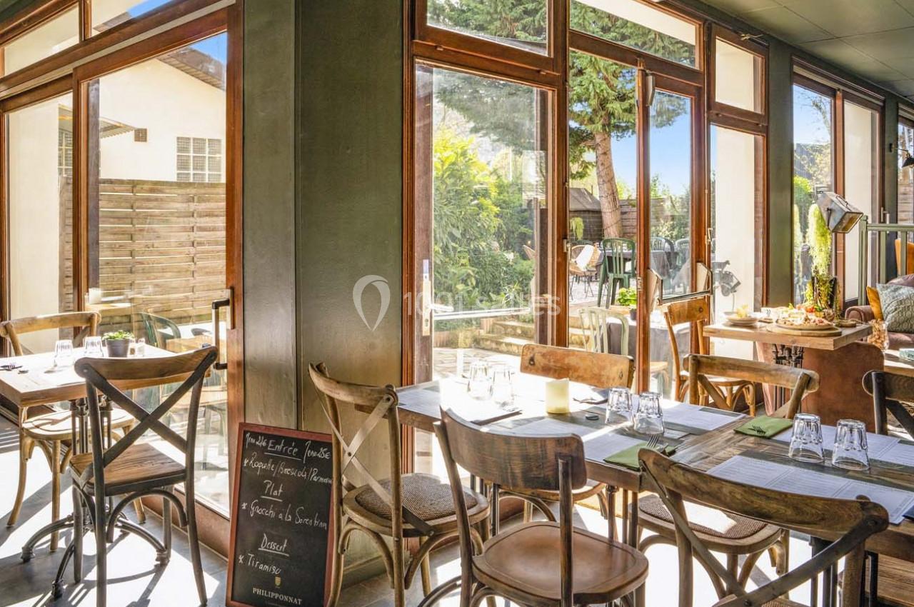 Salle de restaurant lumineuse avec grandes baies vitrées, tables en bois dressées et vue sur un jardin extérieur.