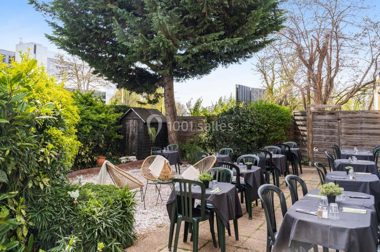 Terrasse extérieure avec tables dressées, chaises noires, plantes verdoyantes et un grand arbre au centre.