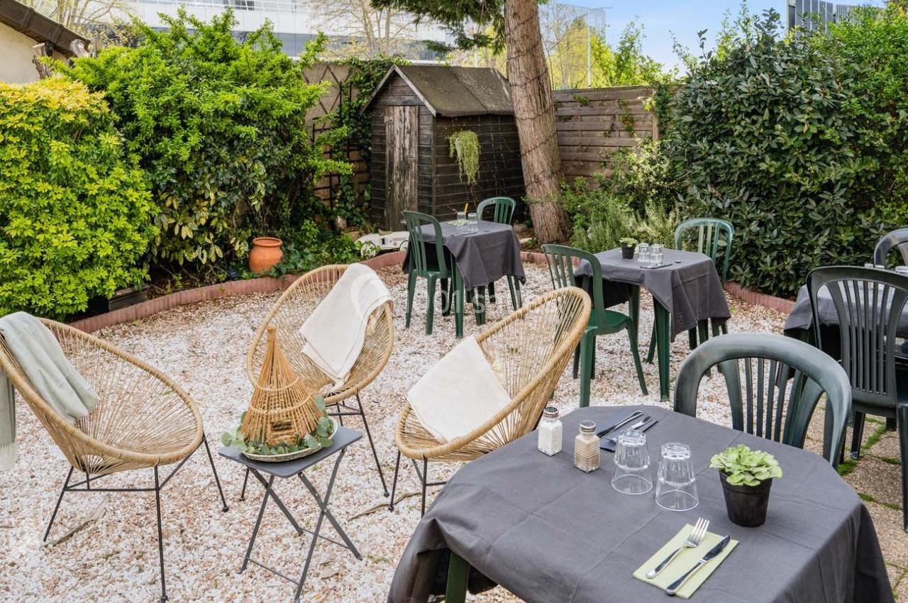 Jardin aménagé avec des tables et chaises pour repas en extérieur, entouré de végétation et d'un cabanon en bois.