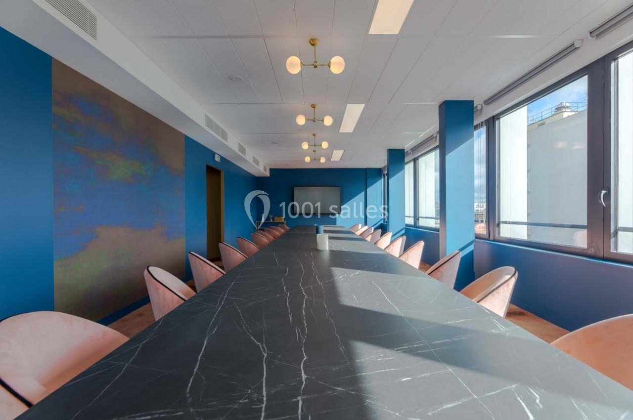 Salle de réunion moderne avec une grande table en marbre noir, chaises roses et murs bleus, éclairée par des fenêtres.