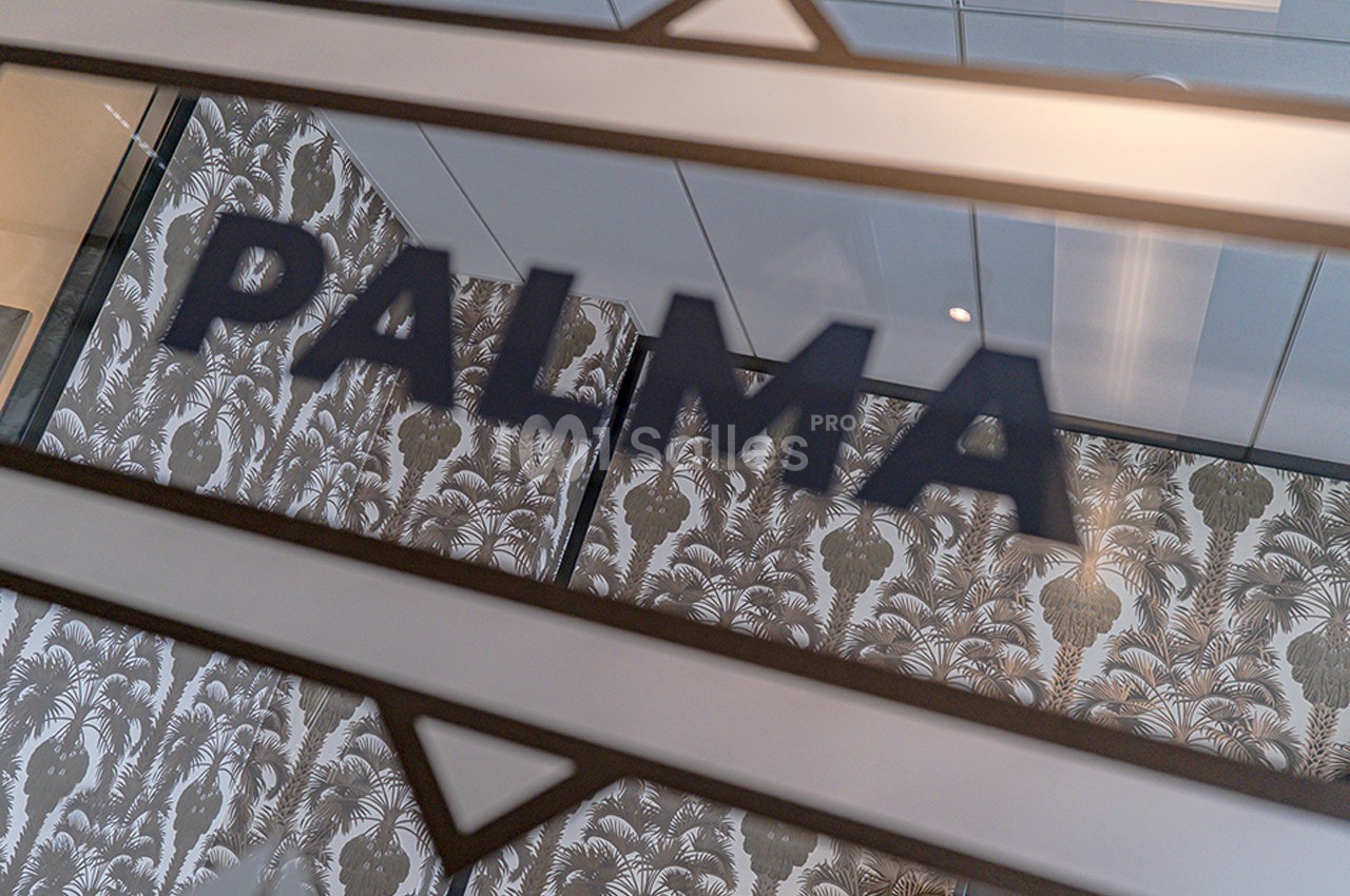 Mot ’PALMA’ inscrit sur une vitre, avec un motif de palmiers en arrière-plan et un plafond moderne visible.