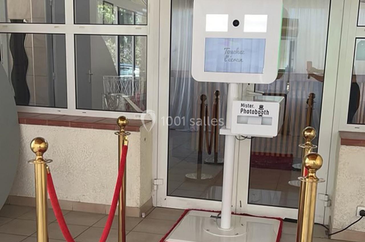 Borne photo interactive blanche installée devant une entrée vitrée, entourée de poteaux dorés et d'un cordon rouge.