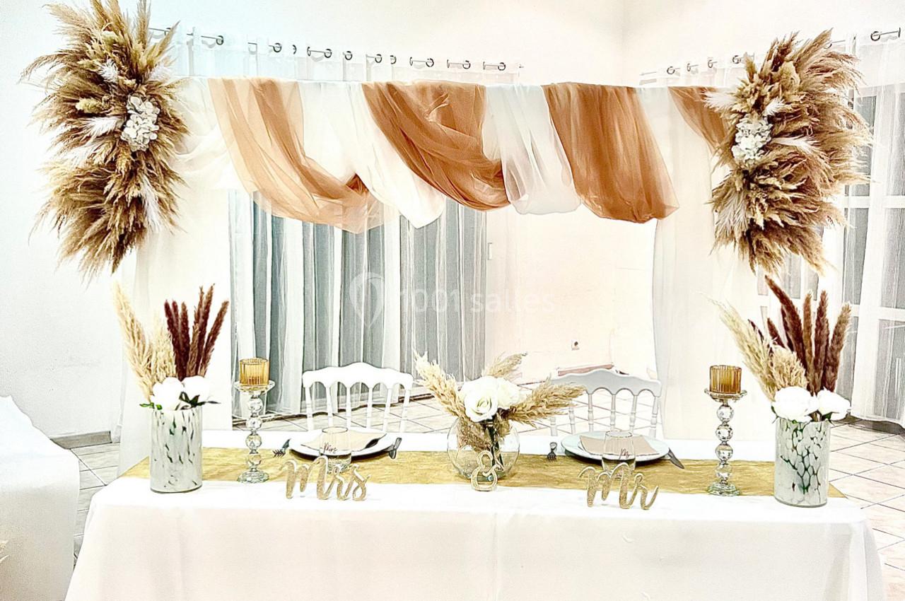 Table décorée pour un mariage avec des fleurs séchées, des voilages beige et blanc, et des chaises blanches en arrière-plan.
