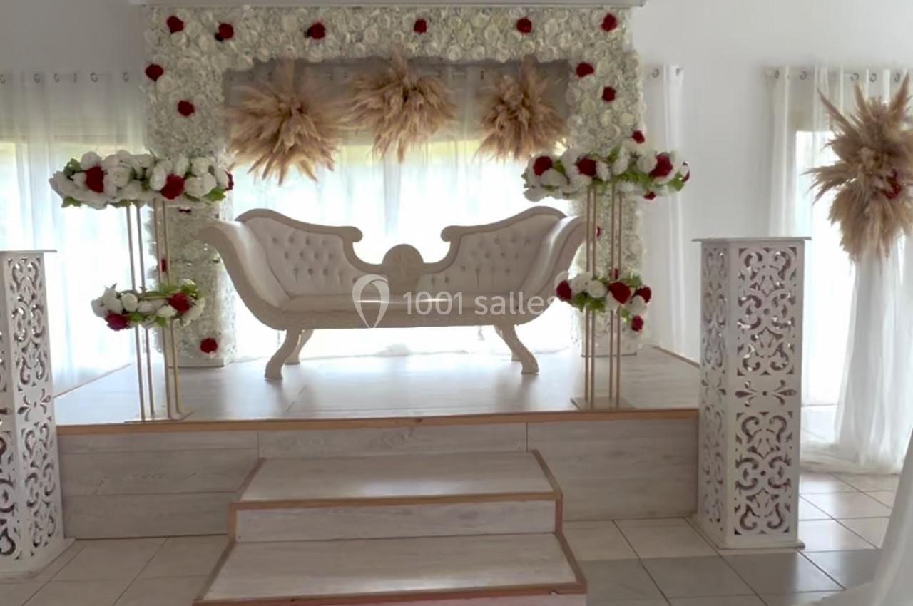 Salle décorée pour un événement avec un canapé blanc sur une estrade, fleurs rouges et blanches, et tapis rouge au sol.