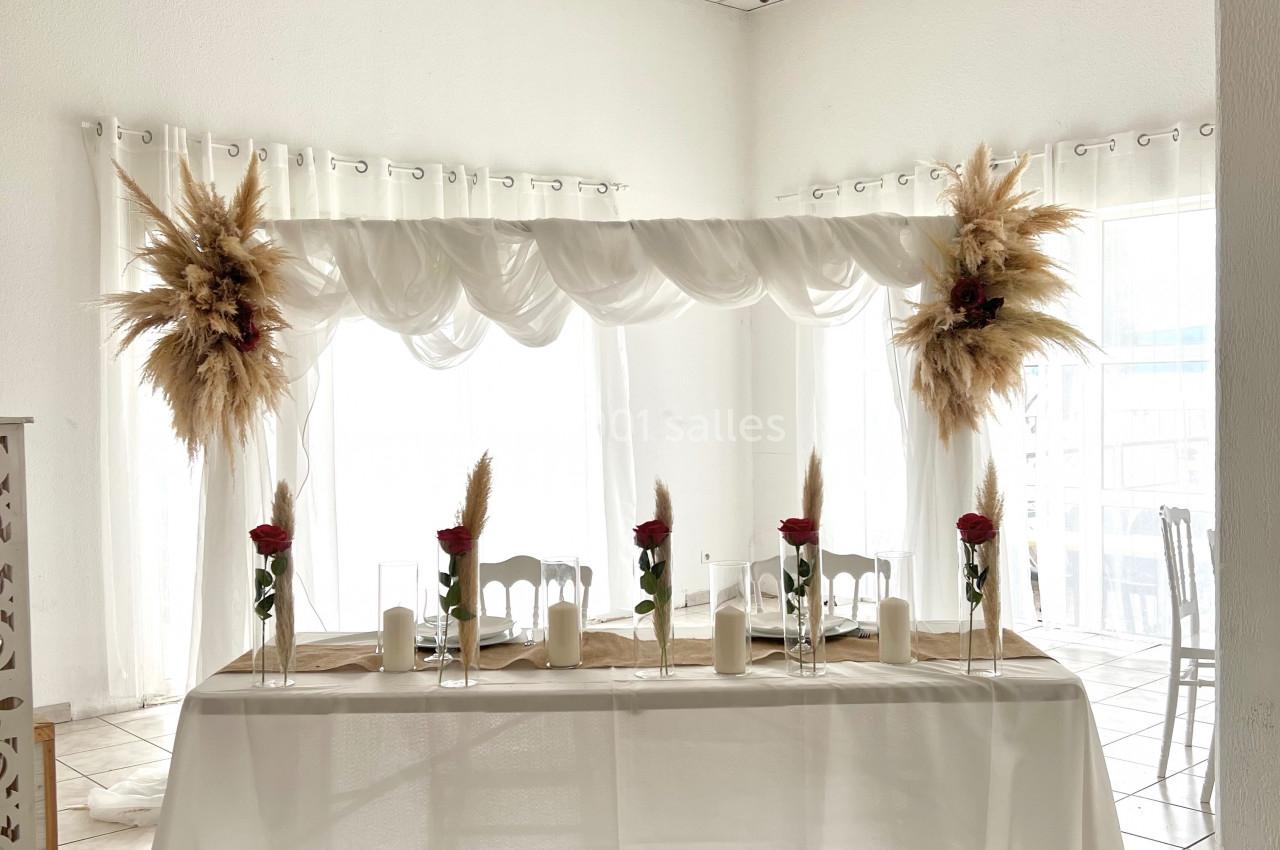 Table décorée pour un événement avec nappes blanches, bougies, fleurs et ornements de pampas dans une salle lumineuse.