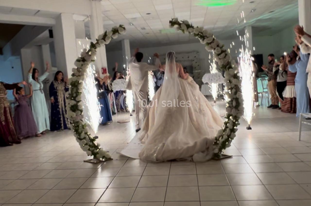 Un couple de mariés traverse une arche fleurie illuminée par des étincelles, entouré d'invités dans une salle de réception.