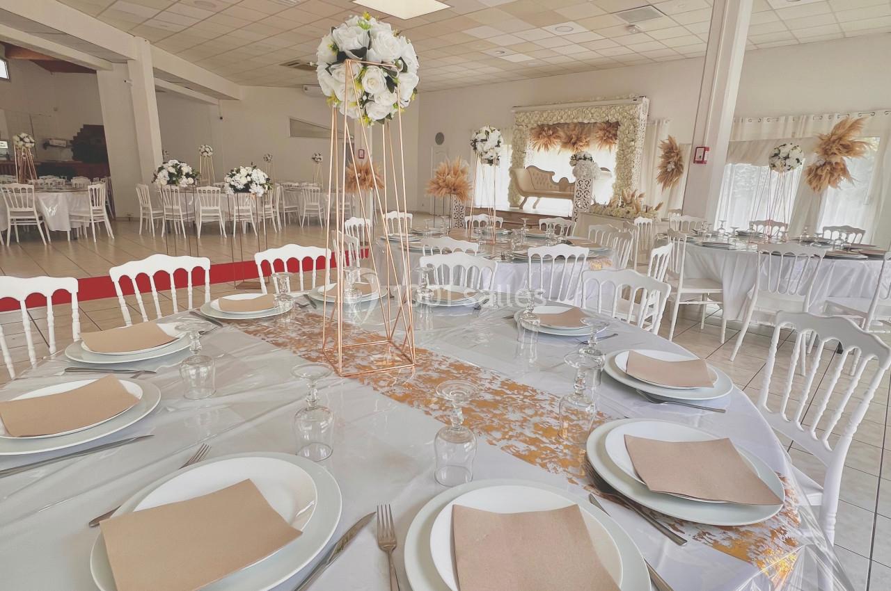 Salle de réception décorée avec des tables dressées, nappes blanches, serviettes beige et centres de table floraux.