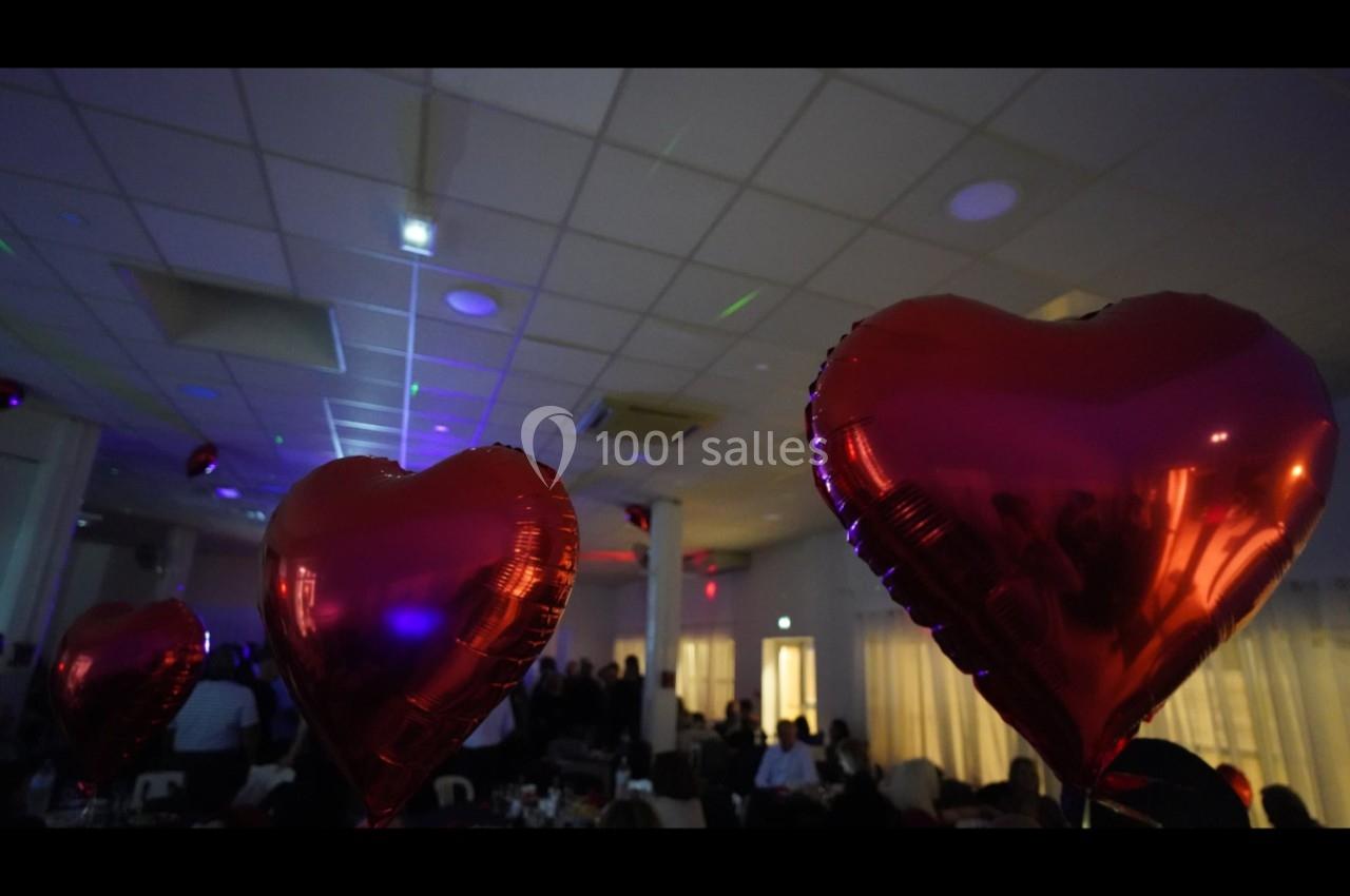 Des ballons rouges en forme de cœur flottent dans une salle éclairée, décorée pour un événement festif.