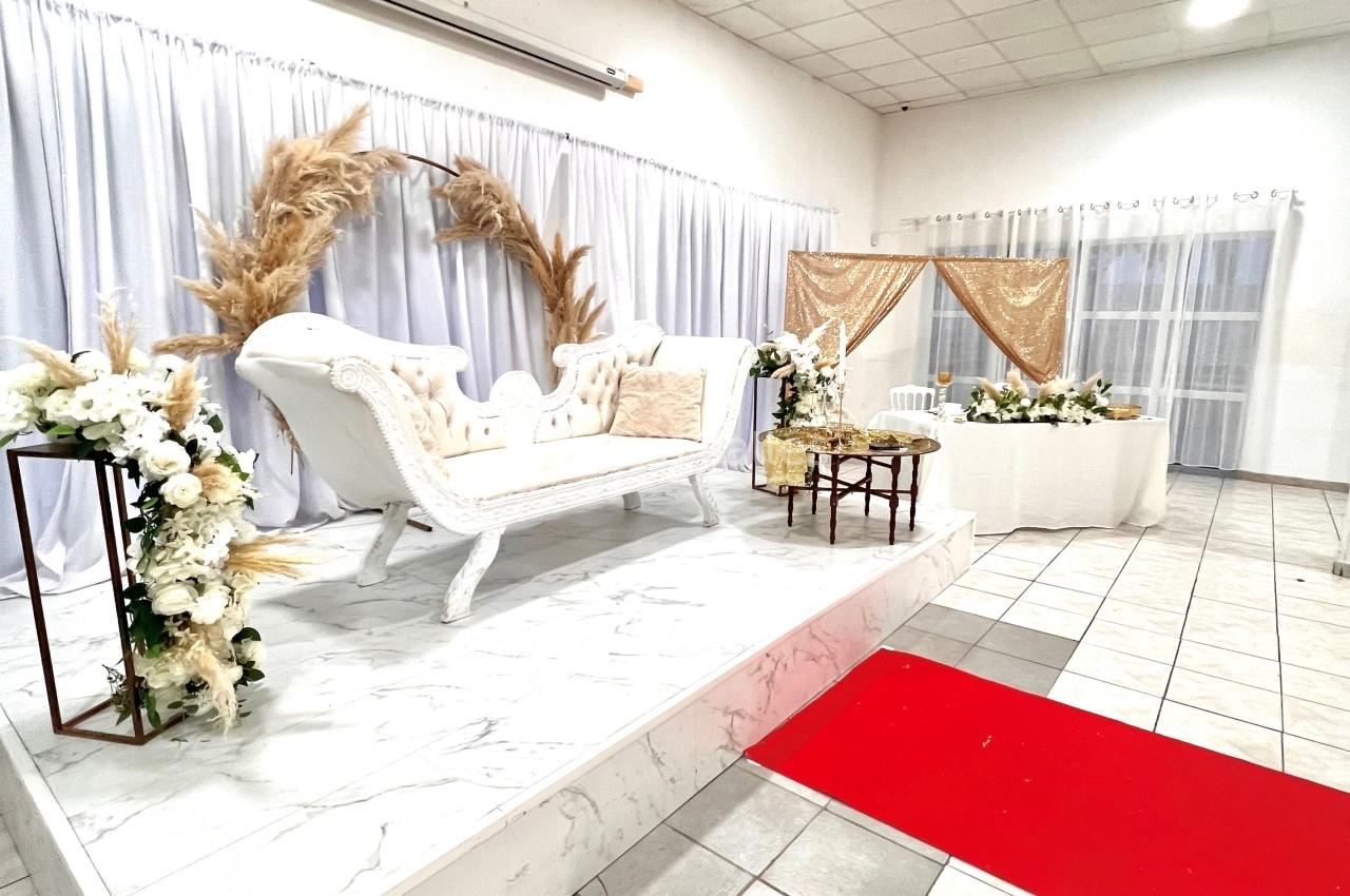 Salle décorée pour un événement avec canapé blanc sur estrade, fleurs, rideaux blancs et tapis rouge au sol.