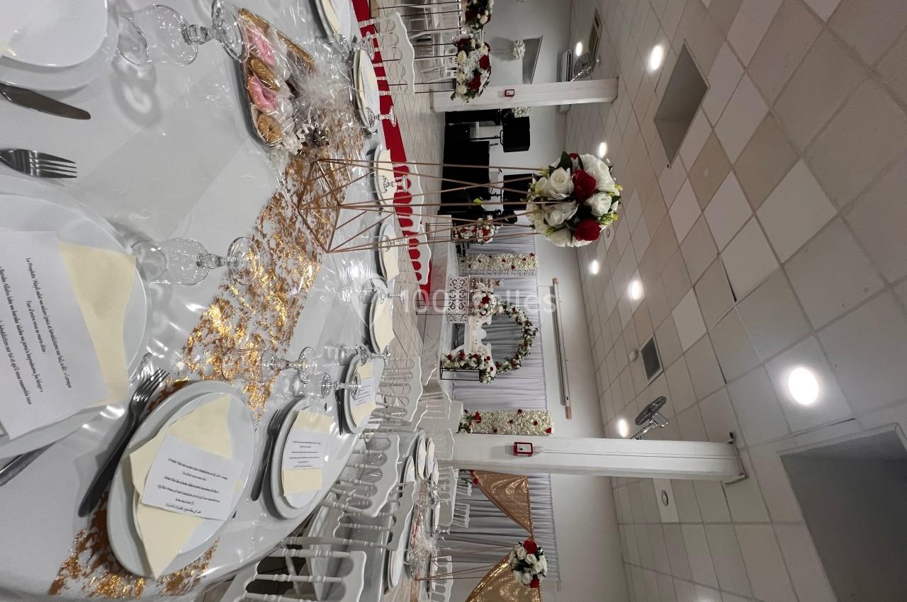 Salle de réception décorée pour un mariage, avec tables dressées, fleurs blanches et rouges, et arche florale en arrière…