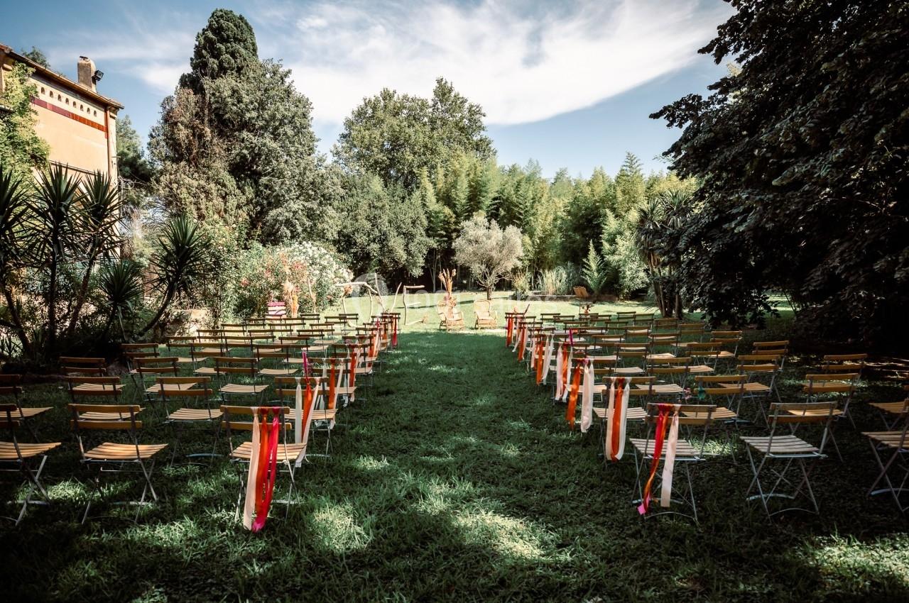 Chaises disposées en rangées sur une pelouse, formant une allée centrale, dans un jardin arboré sous un ciel dégagé.