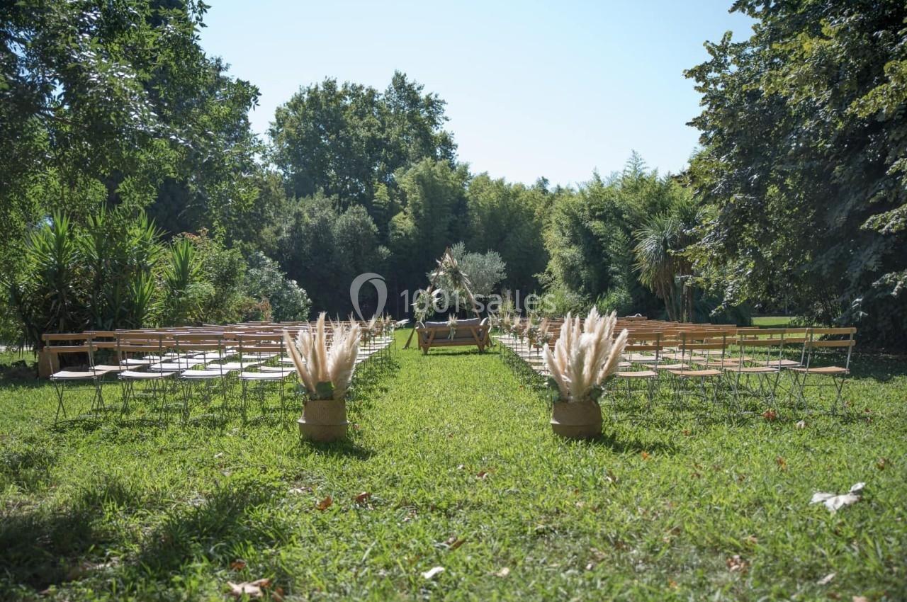Chaises alignées en extérieur, formant une allée centrale décorée de bouquets de pampas, entourée de verdure.