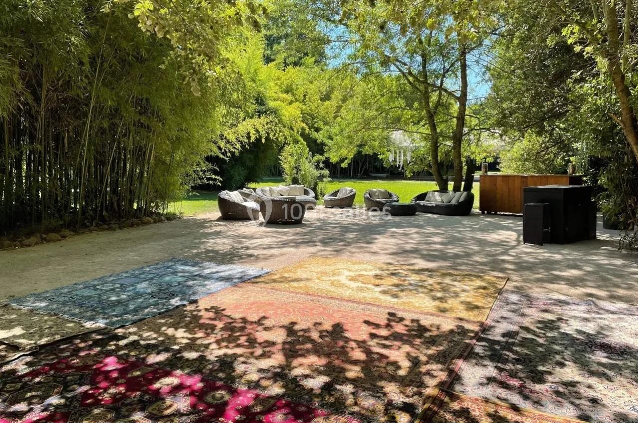 Espace extérieur ombragé avec tapis colorés au sol, fauteuils et végétation luxuriante en arrière-plan.