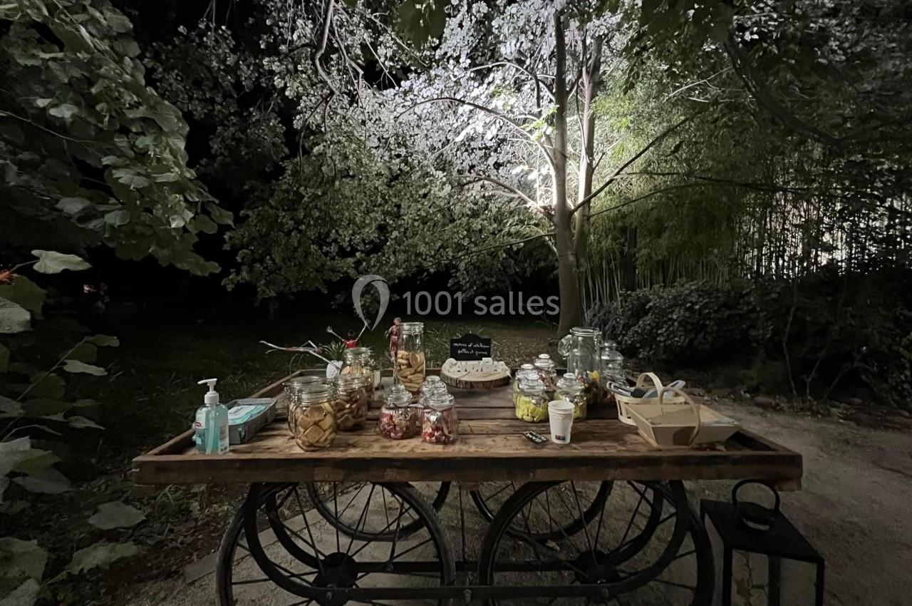 Table en bois sur roues, disposée en extérieur nocturne, garnie de bocaux de friandises et d'objets divers sous un arbre…