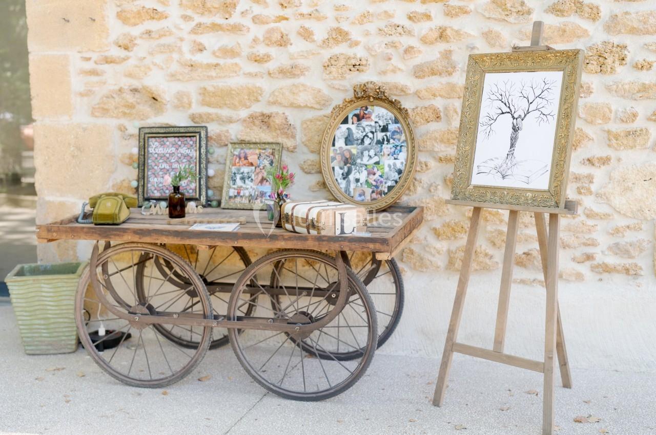 Table en bois sur roues avec cadres photo, fleurs et livres, près d'un chevalet avec un dessin d'arbre, devant un mur en…