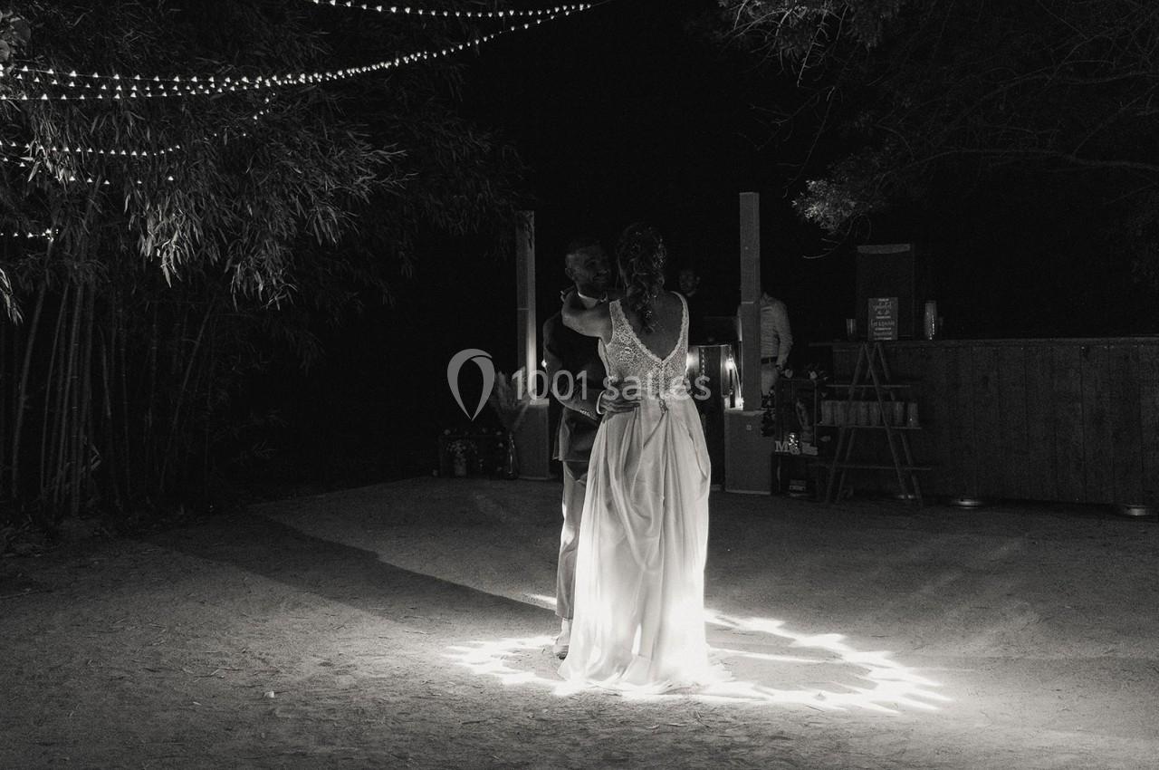 Une mariée en robe blanche se tient sous des guirlandes lumineuses dans un espace extérieur sombre.