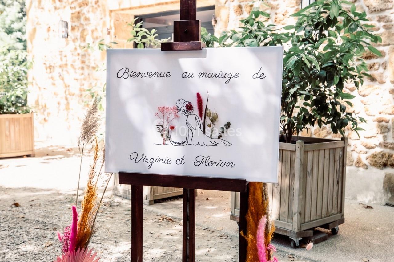 Panneau de bienvenue à un mariage, décoré de fleurs séchées, placé devant un mur en pierre et des plantes.