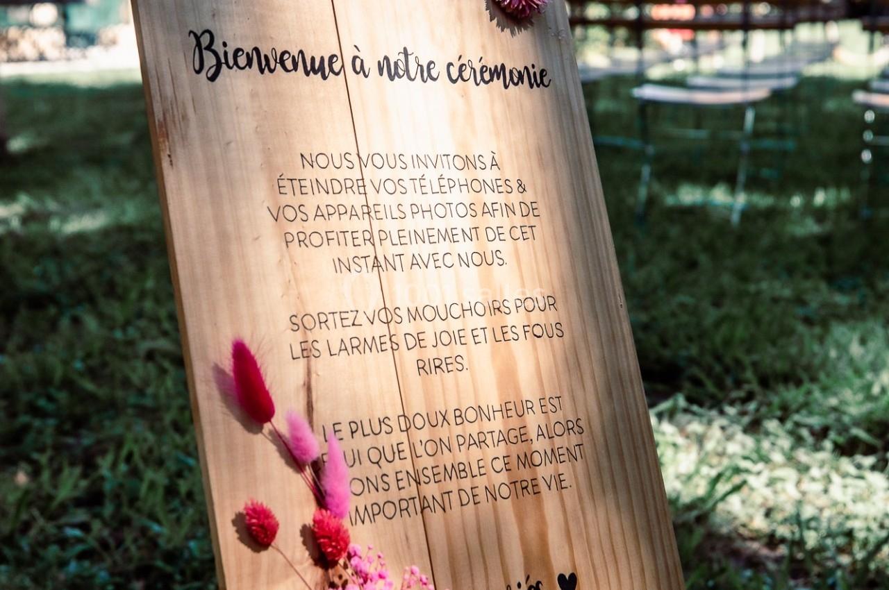 Panneau en bois sur un chevalet, décoré de fleurs séchées, avec un message de bienvenue pour une cérémonie en extérieur.