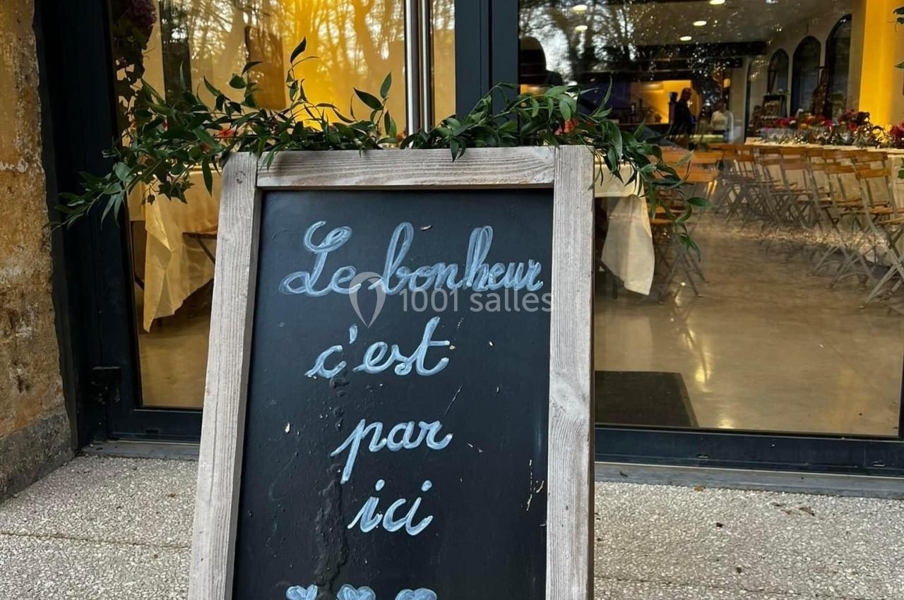 Tableau noir devant une vitrine, inscrit ’Le bonheur c'est par ici’, entouré de feuillage décoratif.