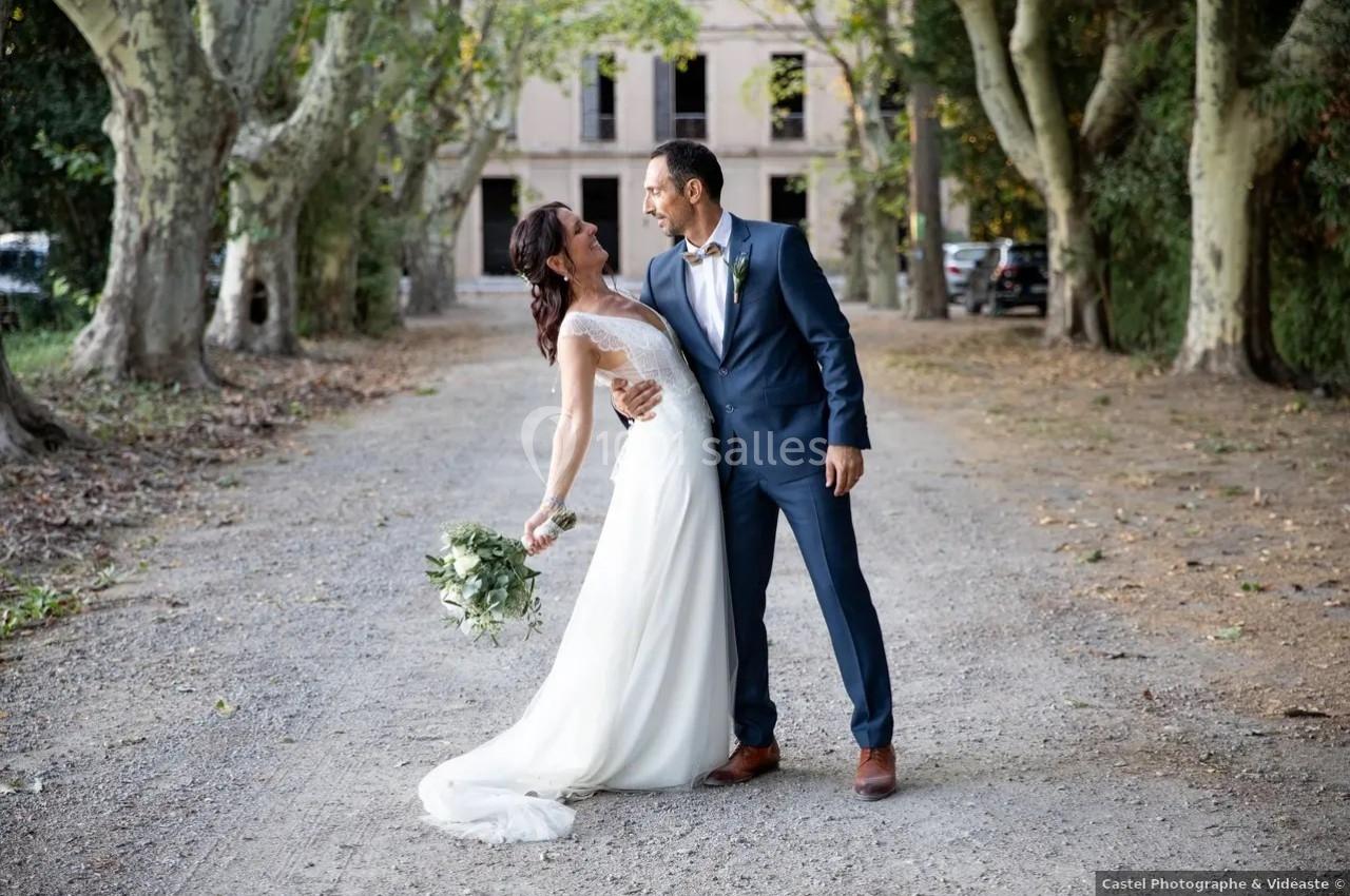 Un couple de mariés souriants pose sur une allée bordée d'arbres devant un bâtiment ancien.
