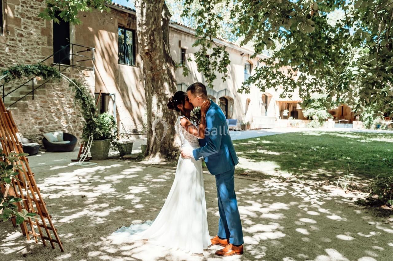 Un couple en tenue de mariage s'embrasse dans un jardin ombragé devant un bâtiment en pierre.