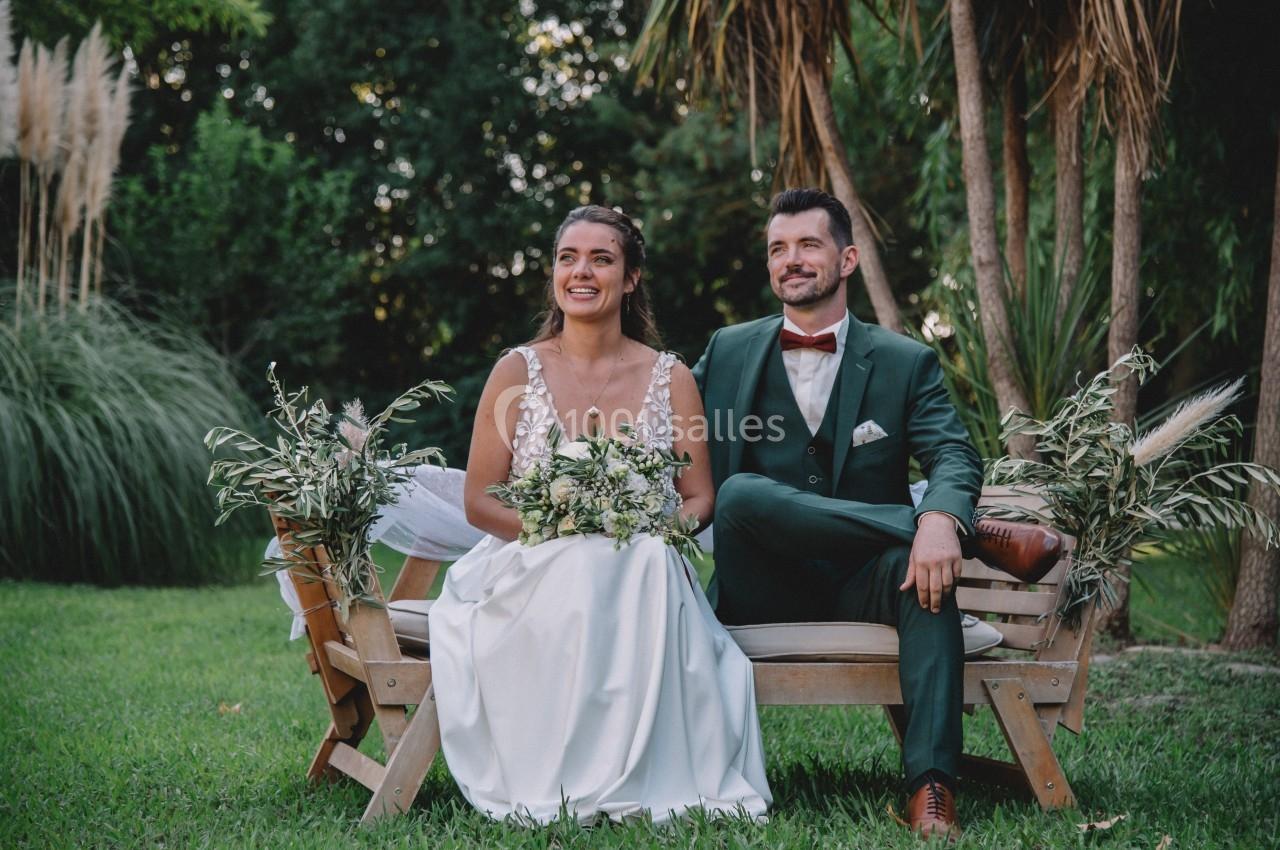 Un couple en tenue de mariage est assis sur un banc en bois dans un jardin verdoyant.