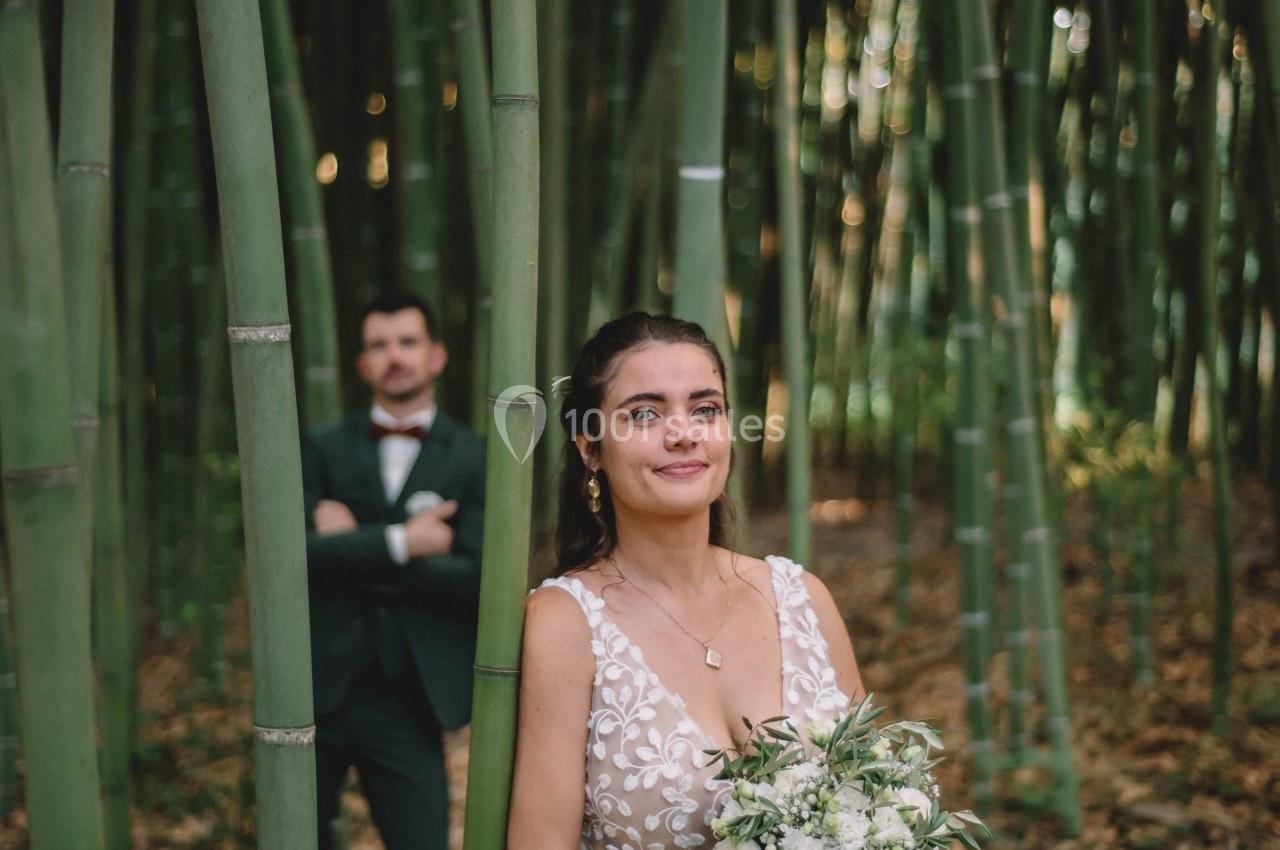 Une femme en robe blanche tient un bouquet dans une forêt de bambous, un homme en costume se tient en arrière-plan.