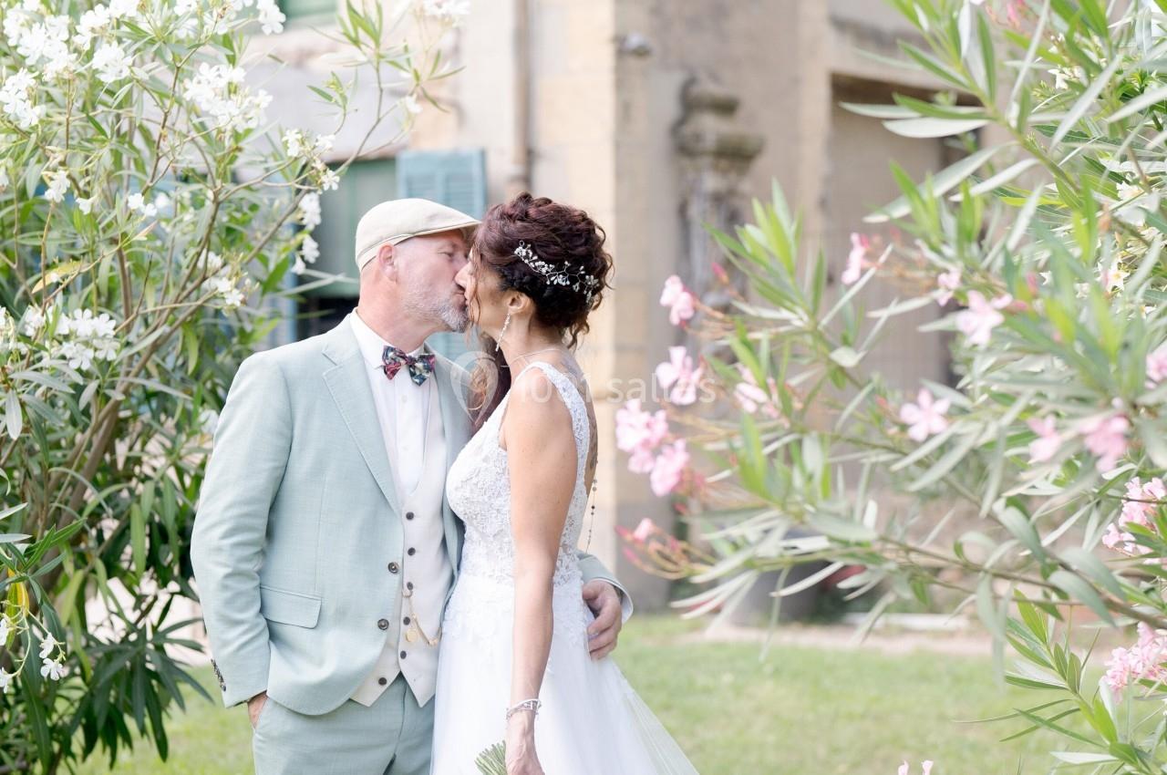 Un couple de mariés s'embrassant dans un jardin, entouré de lauriers roses et d'un bâtiment ancien en arrière-plan.