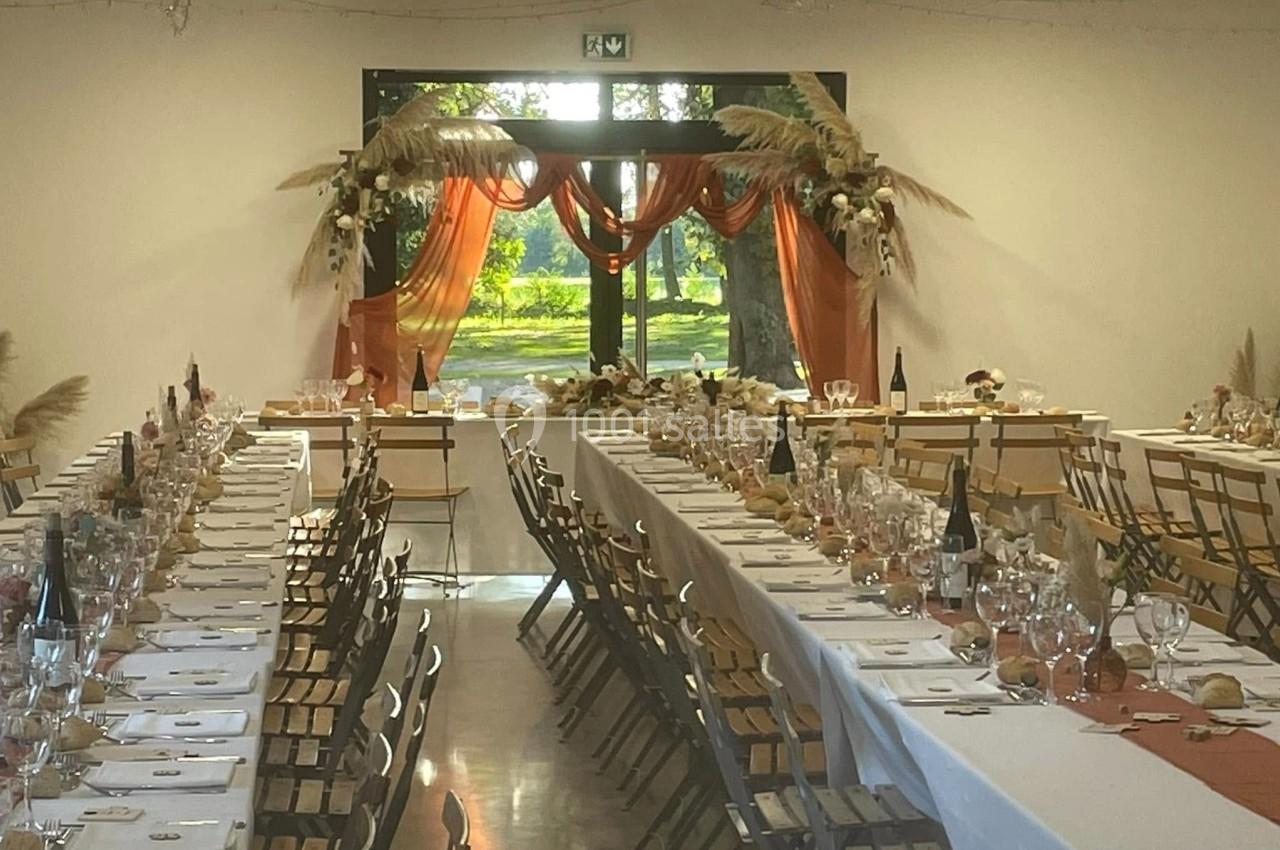 Salle décorée pour un événement avec longues tables dressées, nappes blanches et arrangements floraux près d'une baie vitrée.
