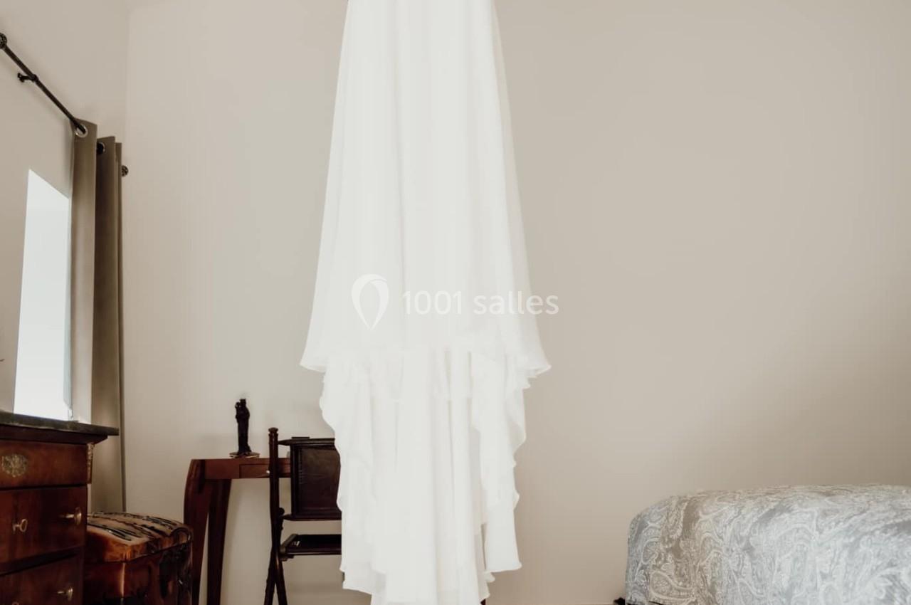 Robe de mariée blanche suspendue à une poutre en bois dans une chambre lumineuse, avec des chaussures posées au sol.