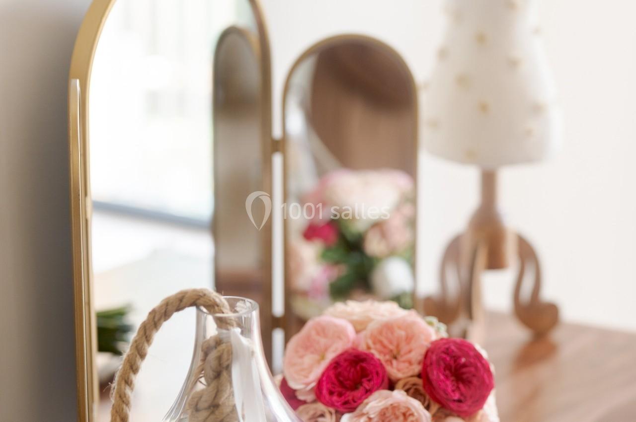 Un vase en verre avec des fleurs blanches, un bouquet de roses et un mannequin décoratif sur une table en bois.